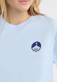 Jasnoniebieska bawełniana koszulka z okrągłą naszywką z haftem przedstawiającą górski motyw oraz tekstem „Ski Club” w kolorach czerwonym, białym i niebieskim.