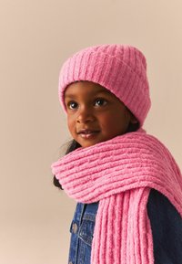 Roze gebreide beanie en bijpassende sjaal met ribbeltextuur. De sjaal is om de nek gewikkeld, gecombineerd met een denimjack.