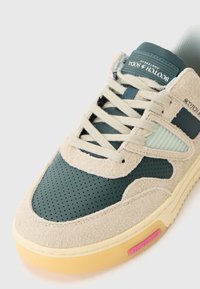 Baskets en daim vert et cuir perforé avec des accents beiges, lacets plats beiges, et une semelle en caoutchouc jaune portant une étiquette de logo rose.