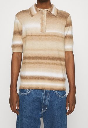 Mężczyzna w krótkim rękawie dzianinowej koszulce polo w beżowo-białe poziome paski oraz niebieskich dżinsach, stojący na jednolitym tle.