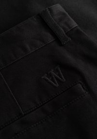 Pantalon noir avec une texture lisse et mate, doté d'une poche cousue. Un logo "W" brodé est discrètement intégré sur la poche arrière.