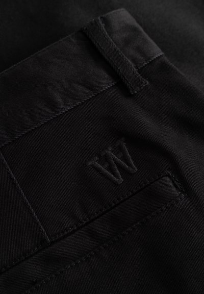 Pantalon noir avec une texture lisse et mate, doté d'une poche cousue. Un logo "W" brodé est discrètement intégré sur la poche arrière.