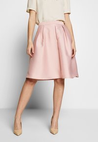 Falda de talle midi en rosa pálido con pliegues, hecha de tela suave. Combinada con tacones beiges de punta. Diseño de cinturilla simple.