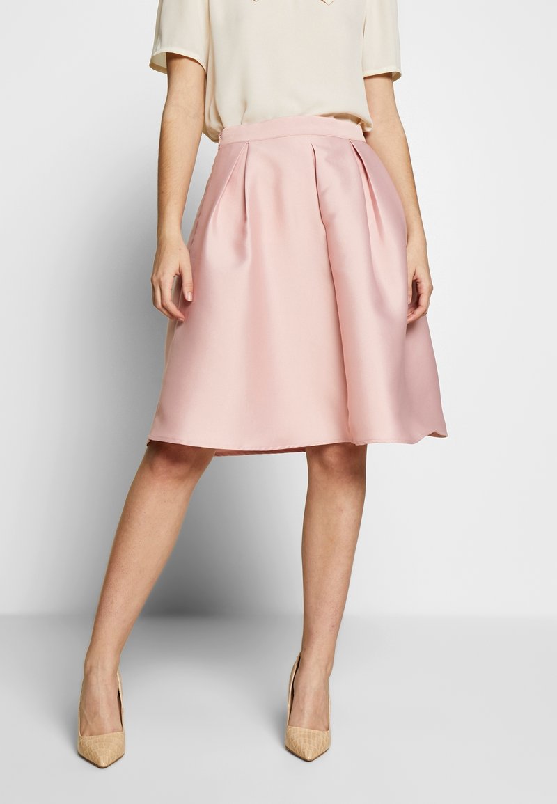 Falda de talle midi en rosa pálido con pliegues, hecha de tela suave. Combinada con tacones beiges de punta. Diseño de cinturilla simple.