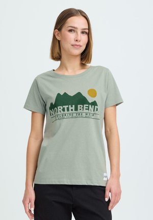 Kvinde med glat brunt hår iført lysegrøn t-shirt med bjerg, sol og teksten "North Bend Exploring The Wild" samt sorte bukser.