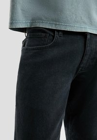 Zbliżenie na osobę ubrana w czarne dżinsy i wyblakły niebieski t-shirt na jednolitym jasnym tle.