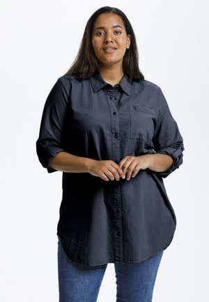 Ung kvinde med langt mørkt hår iført en løs mørk skjorte med knapper og blå jeans står mod en ensfarvet hvid baggrund.