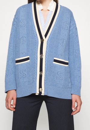 Lichtblauw gebreid vest met zwarte en crème gestreepte rand, zilveren knopen, steekzakken aan de voorkant, gedragen over een gestreept shirt en pantalon met krijtstreep.