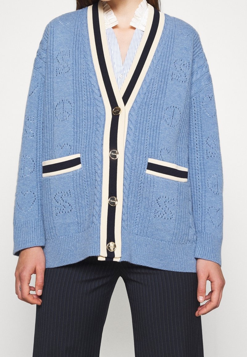 Cardigan bleu clair en maille avec bordure rayée noire et crème, boutons argentés, poches avant, porté sur une chemise rayée et un pantalon à fines rayures.