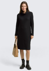 Robe en maille noire à col bénitier, manches longues et silhouette droite, assortie à un sac à main beige et des bottines noires.