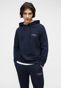 Νεαρός με μεσαίου μήκους μαλλιά, φοράει μπλε σκούρο φούτερ με κουκούλα και αντίστοιχο παντελόνι φόρμας με λογότυπα "Jack & Jones Archive", στέκεται με τα χέρια στις τσέπες.