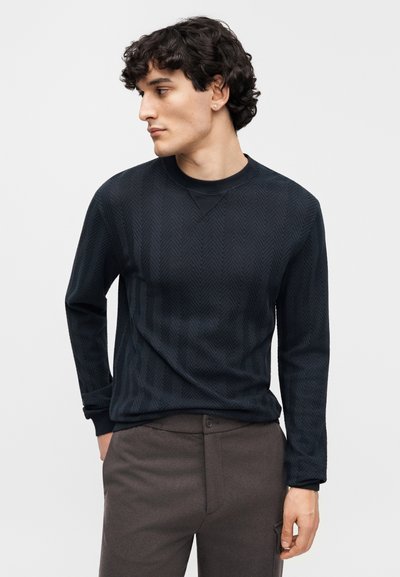 Μαύρο πουκάμισο με μακριά μανίκια και σχέδιο ψαροκόκκαλο, με στρογγυλή λαιμόκοψη και υφασμάτινη υφή, συνδυασμένο με καφέ παντελόνια.