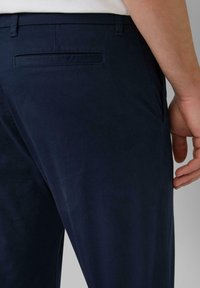 Detailný záber osoby, ktorá má na sebe tmavomodré nohavice s vreckom na zadnej strane a viditeľnou rukou odpočívajúcou pri bokoch.