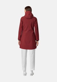 Cappotto impermeabile rosso con cappuccio, con maniche lunghe e taglio dritto. Indossato sopra pantaloni bianchi e calzature bianche. Finitura opaca liscia.