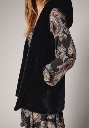 Femme portant un gilet sans manches en fourrure noire sur une robe à manches longues à motif cachemire avec des accents bleus, orange et beige.