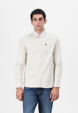 Chemise beige clair à manches longues avec un col boutonné et une poche poitrine, ornée d'un logo brodé jaune ; portée avec un jean bleu.