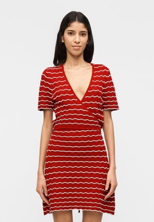 Γυναίκα ντυμένη με κόκκινο πλεκτό φόρεμα κοντών μανικιών με λευκές οριζόντιες κυματοειδείς ρίγες και βαθύ V-λαιμόκοψη σε απλό φόντο.