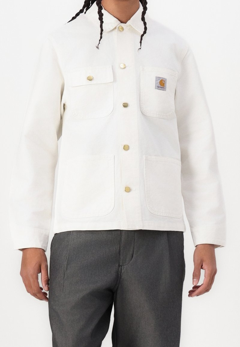 Persona vestida con una chaqueta blanca de botones y cuatro bolsillos frontales, con un parche del logo de Carhartt en el bolsillo del pecho, combinada con pantalones gris oscuro.