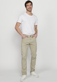 Svetlo béžové džínsy s piatimi vreckami, slim fit, zapínanie na päť gombíkov. Nosené s jednoduchým bielym tričkom a bielymi teniskami.