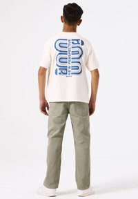 Wit katoenen t-shirt met een blauw grafisch ontwerp op de achterkant, gecombineerd met lichtgroene broeken en witte sneakers.