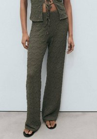 Olivovo zelený cropped top a široké nohavice z textúrovaného materiálu. Voľný strih so šnúrkou v páse. Čierne sandále na otvorenej špičke.