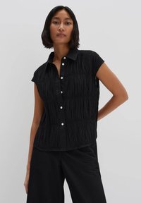 Maglietta nera senza maniche con pieghe verticali testurizzate, abbottonatura frontale e colletto. Realizzata in un tessuto leggero.