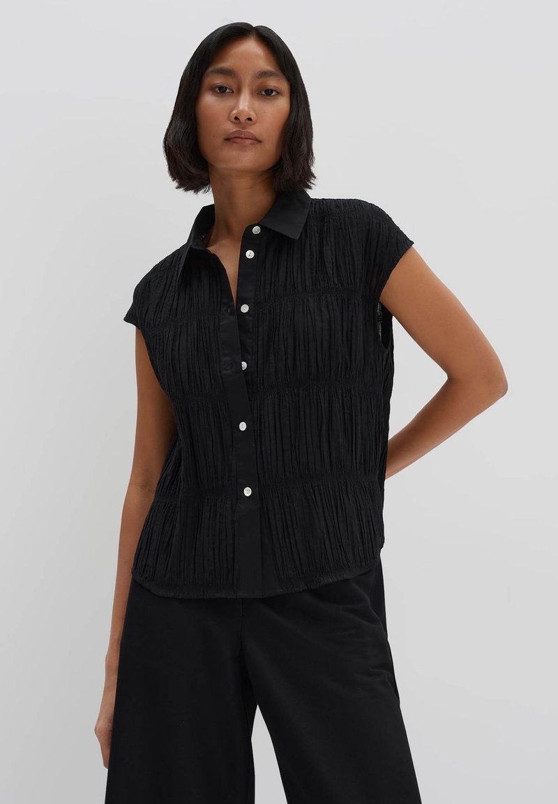 Maglietta nera senza maniche con pieghe verticali testurizzate, abbottonatura frontale e colletto. Realizzata in un tessuto leggero.