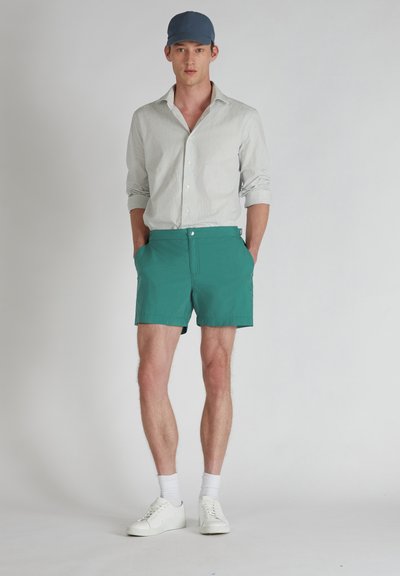 Bañador verde con bolsillos, combinado con una camisa de rayas blanca y azul, zapatillas blancas y una gorra azul. Textura suave, ajuste casual.