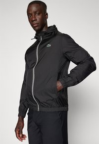 Zwarte lichtgewicht zip-up jas met capuchon, elastische manchetten en tailleband. Bevat een klein groen logo op de borst en zijzakken.