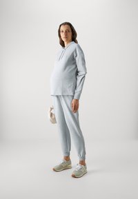 Svetlo šedá mikina s kapucňou a zodpovedajúce jogger tepláky z jemnej látky, s voľným strihom a elastickými manžetami, v kombinácii so zelenými a bielymi teniskami.