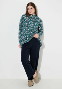 Monivärinen paita kukkakuviolla, korkea kaulus, pitkät hihat. Tummansiniset kuvioidut housut, beigekudotut kengät.