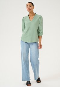 Blouse vert clair avec un col volanté, manches bouffantes trois-quarts, associée à un jean bleu ample et des sandales noires à bout ouvert.
