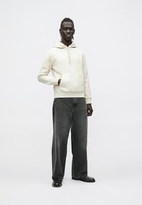 Kremna jopa s kapuco, s sprednjim žepom in toniranim logotipom, kombinirana s širokimi temno sivimi kavbojkami. Moški stoji sproščeno, obut v črne čevlje.