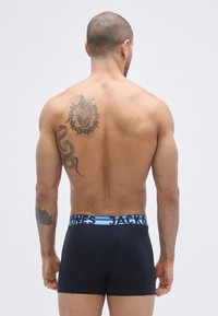 Mustat bokserishortsit, joiden vyötäröllä on sinimusta vyö, jossa lukee "JACK&JONES"; mallilla on näkyvä tatuointi selässään.