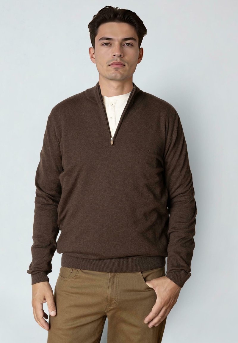 Nuori mies, joka on pukeutunut ruskeaan neljännesvetoketjulliseen villapaitaan valkoisen paidan ja ruskeiden housujen päällä, seisoo toinen käsi taskussa vaaleaa yksinkertaista taustaa vasten.