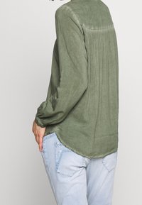 Camicia a maniche lunghe verde oliva con schiena arricciata, orlo arrotondato e tessuto testurizzato. Abbinata a jeans azzurri chiari.