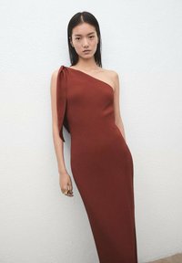 Bordowa sukienka na jedno ramię z drapowanym lewym rękawem, dopasowanym krojem i gładką fakturą, zestawiona ze złotym pierścieniem na dłoni modelki.