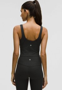 Sort atletisk top med en tætsiddende design, brede stropper, diskret tekstureret mønster og logo detalje på ryggen. Bæres med matchende leggings.