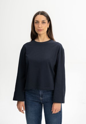 Vrouw met lang donker haar, gekleed in een donker langärmelig shirt en een blauwe spijkerbroek, staand tegen een effen witte achtergrond.
