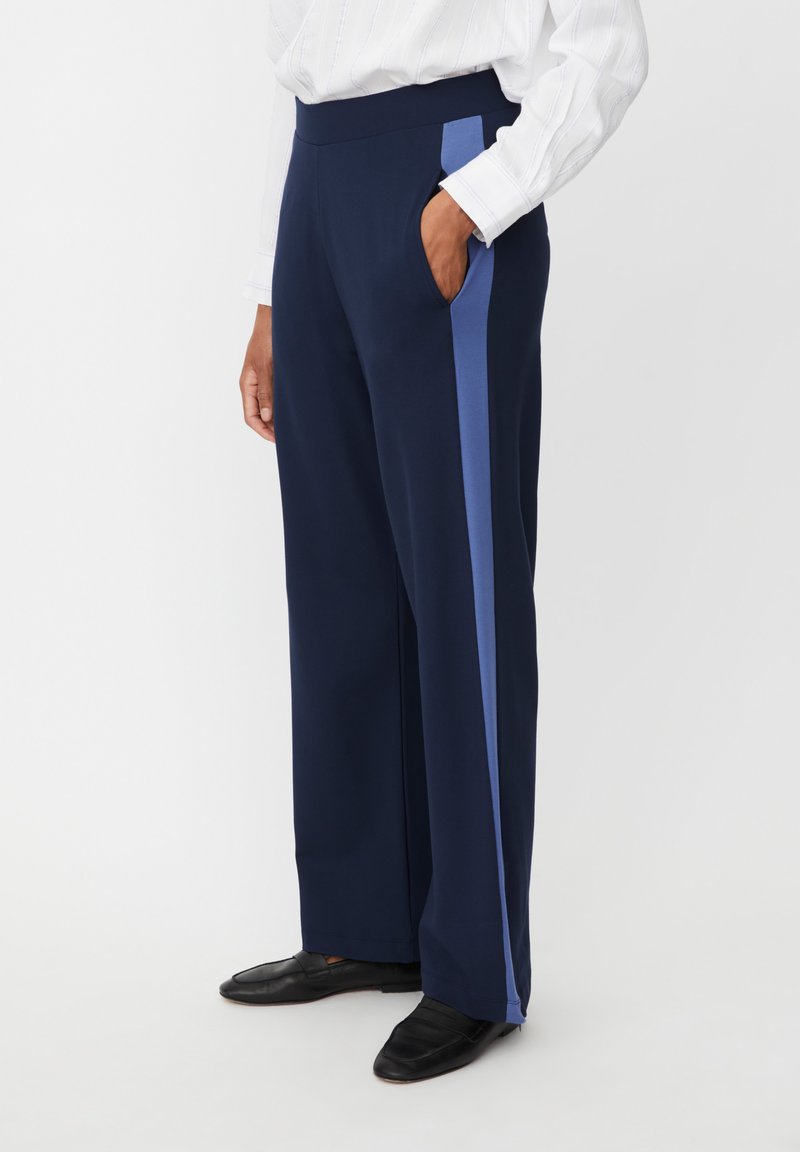 Granatowe spodnie o szerokich nogawkach z jasnoniebieskimi bocznymi paskami, gładką tkaniną, elastycznym pasem i bocznymi kieszeniami. Noszone z czarnymi mokasynami.