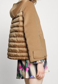 Padded brauner Mantel mit einer strukturierten Wolloberfläche, ausgestattet mit einer Kapuze und großen Taschen, kombiniert mit einem rock mit floralem Muster.