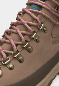 Κοντινό πλάνο μπεζ πεζοπορικού μποτάκι με κορδόνια μπεζ με ροζ λεπτομέρειες περασμένα μέσα από μεταλλικά μάτια D-ring σε ταμπά σουέτ επάνω μέρος.