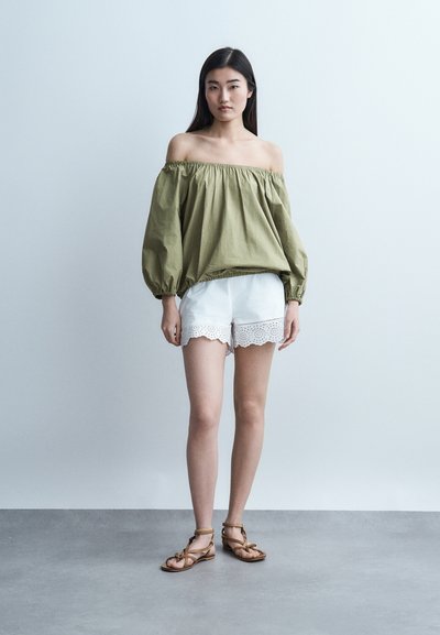 Νεαρή γυναίκα όρθια, ντυμένη με χακί μπλούζα με έξω τους ώμους, λευκά σορτς με δαντέλα eyelet και καφέ φλατ σανδάλια με λουράκια, σε γκρι πάτωμα.