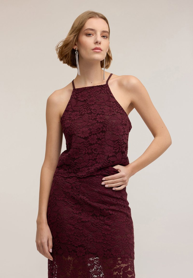 Vinröd spetsklänning med halterneck, figursydd överdel och flytande kjol. Detaljerat blommönster och sidofickor med utskärningar.