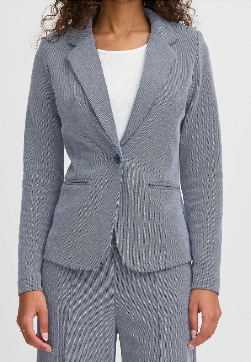Personne portant un blazer gris ajusté avec un seul bouton et un pantalon assorti sur un haut blanc côtelé.