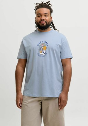 Muškarac s bradom i dredovima nosi svijetloplavu Jack & Jones majicu s motivom lubanje i palme te bež hlače, stoji na bijeloj pozadini.