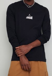 Mustat pitkähihainen t-paita, jossa on valkoinen "JORDAN" -logo rinnassa. Paita on valmistettu puuvillasta, ja siinä on istuvat hihansuut ja pyöreä kaula-aukko.
