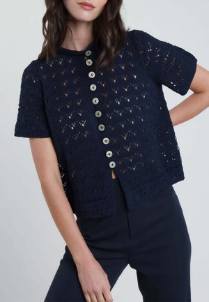 Donna che indossa un cardigan corto in crochet blu navy con bottoni simili a perle sul davanti e pantaloni blu navy abbinati.