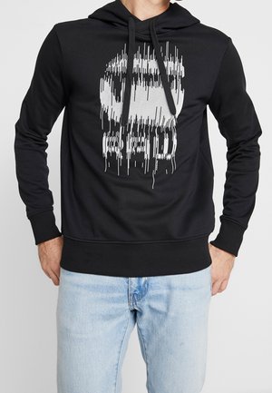 Man draagt een zwarte hoodie met een witte vervormde schedel en de tekst "RAD", gecombineerd met lichtblauwe spijkerbroek, staand met handen in de zakken.