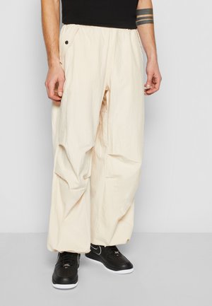 Muž v pohodlných voľných nohaviciach off-white s širokými nohavicami a postrannými vreckami na gombíky, v čiernych teniskách s bielymi podrážkami na jednoduchom pozadí.